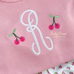 May include: A pink shirt with a white embroidered letter R and two cherries. The shirt is paired with white pants with a cherry print.