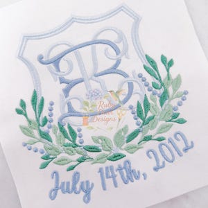 May include: White fabric with a light blue embroidered design. The design includes a monogram with the letters "RB" inside a shield, surrounded by a green and blue floral wreath. The text "July 14th, 2012" is embroidered below the wreath.