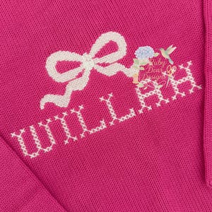 Puede incluir: Un conjunto de bebé de punto magenta con el nombre "WILLAH" bordado en punto de cruz blanco. El diseño incluye un lazo blanco, flores y un colibrí. El texto "Baby Dear Designs" también es visible.