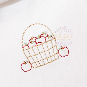 May include: A white embroidered design of a basket filled with red apples and two apples on the side. The basket is brown and has a gold handle.