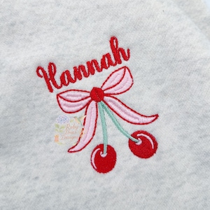 May include: A light gray sweatshirt with the name "Hannah" embroidered in red script above a pink bow and two red cherries with green stems. The design is centered on the garment.
