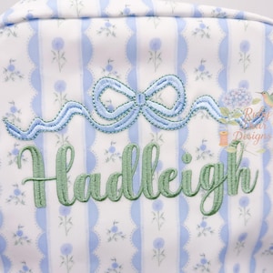 May include: A white and blue floral patterned backpack with a light blue bow and the name "Hadleigh" embroidered in green. The backpack has a scalloped edge design and a zipper closure.