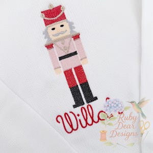 Könnte beinhalten: Weißes Strickkleidungsstück mit einem gestickten Nussknackersoldaten in Rosa, Rot und Schwarz. Der Name "Willow" ist in roter Schrift unter dem Nussknacker gestickt. Ein kleines Blumen- und Kolibri-Design ist ebenfalls vorhanden.