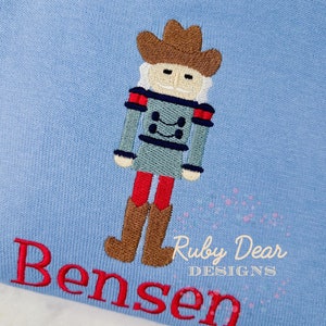 Könnte beinhalten: Ein hellblauer Strickpullover mit einem gestickten Nussknacker-Motiv und dem Namen "Bensen" in roter Stickerei. Der Pullover ist mit einem kleinen "Ruby Dear Designs" Logo verziert.