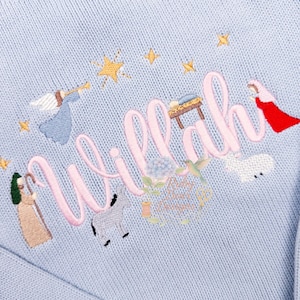 Puede incluir: Un suéter de punto azul claro con la palabra "Willah" bordada en rosa. El diseño incluye imágenes bordadas de estrellas, un ángel, un pastor, un burro, un cordero y una escena del pesebre.
