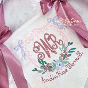 Puede incluir: Manta de bebé bordada blanca con un diseño floral rosa y azul. La manta presenta las iniciales "WR" en una fuente decorativa, rodeadas por una corona floral y el nombre "Sadie Rae Worrell". Una cinta de raso rosa está atada alrededor de la manta.
