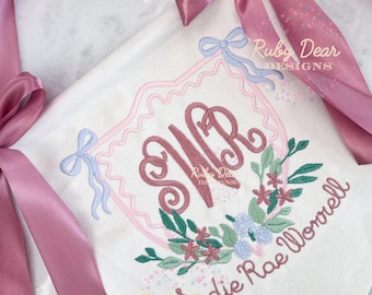 May include: White embroidered baby blanket with a pink and blue floral design. The blanket features the initials "WR" in a decorative font, surrounded by a floral wreath and the name "Sadie Rae Worrell". A pink satin ribbon is tied around the blanket.