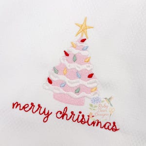 May include: White Christmas-themed hand towel with a pink embroidered Christmas tree, adorned with colorful lights and a yellow star. The words "merry christmas" are embroidered in red. The towel has a decorative, dotted edge.