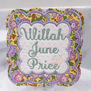 May include: A decorative, scalloped-edge sign with floral fabric and embroidered text. The sign features the names "Ullilah June Price" in green script on a white dotted background, framed by a purple border and a yellow floral pattern.