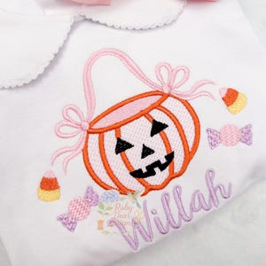 May include: White baby bodysuit with a Peter Pan collar, featuring an embroidered pumpkin basket design in orange and pink, with candy corn and wrapped candy accents. A large pink bow sits at the top. The name "Willah" is embroidered in a cursive font.