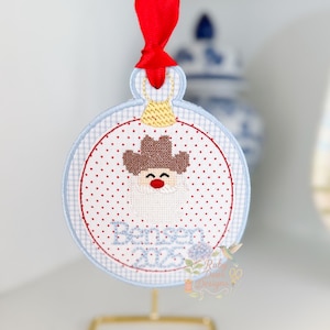 May include: Embroidered Christmas ornament with a cowboy Santa design. The ornament features a red ribbon, a blue and white gingham border, and the text "Better Days 2023". The ornament is displayed on a gold stand.