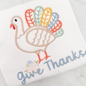 以下が含まれることがあります： ベージュ、オレンジ、黄色、緑、青色の刺繍が施された七面鳥のデザインが施された白いTシャツ。「give thanks」の文字が青色で刺繍されています。感謝祭をテーマにしたデザイン。