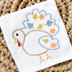May include: White embroidered applique of a turkey with a blue, green, orange, and yellow fall leaf design. The design is on a white t-shirt.