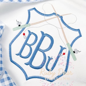Op de afbeelding: Blauw geborduurd monogram met de letters "BB" in een schrijfletter, omringd door twee gekruiste hengels met aas. Het ontwerp is op een witte stoffen achtergrond met een blauw-wit geruit patroon in de hoek.