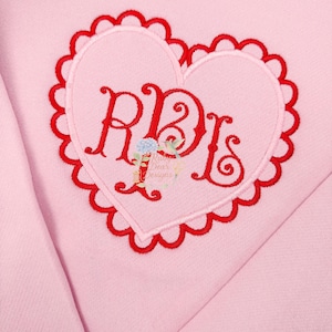 May include: A light pink sweatshirt featuring a red heart-shaped design with a scalloped border. The heart contains the embroidered letters "RBs" in red script, with a small floral accent. The text "Dear Designs" is also visible.