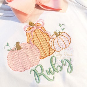 May include: A white onesie with an embroidered design of three pumpkins. The pumpkins are orange, pink, and peach. The top pumpkin has a pink bow. The name "Ruby" is embroidered in green below the pumpkins.