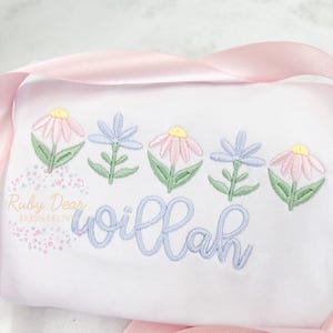 May include: White embroidered baby onesie with a row of five pastel flowers and the name "willah" embroidered in blue thread.