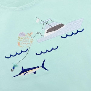 May include: A light mint green t-shirt featuring an embroidered design of a white motorboat on dark blue waves. A fishing line extends from the boat into the water, ending with a hook near a dark blue and white marlin fish swimming below.