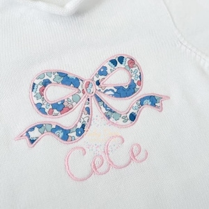 May include: White knit garment featuring a floral bow applique in blue, pink, and green. The bow is outlined in pink, and the name "Cece" is embroidered below in pink script. The garment has a ribbed collar.