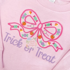 Może przedstawiać: Jasnoróżowa bluza z dużym haftowanym wzorem kokardy. Kokarda wypełniona jest cukierkami, zapakowanymi słodyczami i małymi dyniami. Słowa "Trick or Treat" są wyhaftowane poniżej fioletową czcionką. Bluza z motywem Halloween.