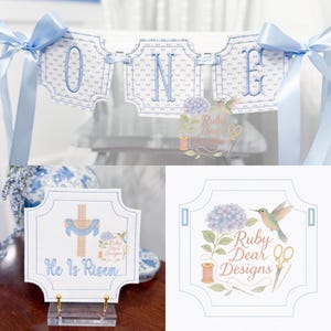 May include: A banner spelling "ONE" in blue embroidered letters, with a matching blue ribbon. Below, a sign with a cross and the text "He Is Risen". The Ruby Dear Designs logo features a hummingbird and flowers.