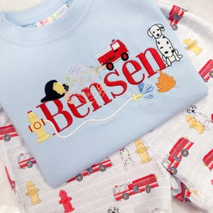 May include: A light blue children's shirt with the name "Bensen" embroidered in red, featuring a fire truck, Dalmatian, and fire-related graphics. The matching pajama pants have a print of fire trucks, fire hydrants, and Dalmatians.
