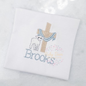 May include: White embroidered design of a lamb standing next to a cross with a blue scarf. The name "Brooks" is embroidered below the design.