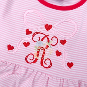 May include: Pink and white striped dress with a red trim around the neckline. The dress features an embroidered bow, red hearts, and the letter "A" in red. The text "Baby Dear Designs" is also embroidered on the dress.