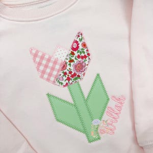 Pode incluir: Um moletom rosa claro com uma aplicação floral. A aplicação inclui um design de tulipa em patchwork com tecidos rosa xadrez, florais e pontilhados. A haste e as folhas são verdes, e a palavra "Tillah" é bordada em rosa.