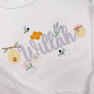 May include: White long-sleeved shirt with the name "Willah" embroidered in lavender script. The design includes bees, a honeycomb, a beehive, and a jar of honey. The shirt has a round neckline and a ruffle detail at the sleeve.