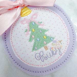 May include: Embroidered Christmas ornament with a pink bow and ribbon. The ornament features a green Christmas tree design with colorful accents and the name "Ruby" stitched in cursive. The ornament has a light purple border.