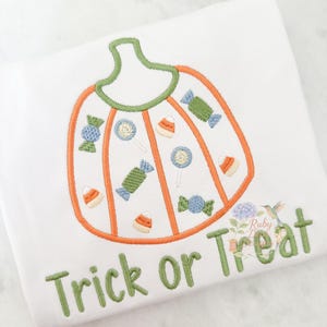 Puede incluir: Camiseta blanca con un diseño de calabaza bordada llena de caramelos y las palabras "Trick or Treat" en verde. El contorno de la calabaza es naranja con un tallo verde.