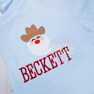 May include: Light blue t-shirt with an embroidered cartoon image of a cowboy wearing a brown hat and a white beard. The name "BECKETT" is embroidered in red below the image.