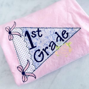 Puede incluir: Camiseta de tela rosa con un parche triangular decorativo. El parche presenta las palabras "1st Grade" en azul marino, con un estampado floral y ribete de lunares. Lazos rosas cosidos en las esquinas.