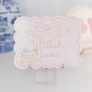 May include: A white, scalloped-edge name card with pink polka dots and a pink bow design. The name "Willah June" is embroidered in pink script. A small floral and hummingbird illustration is in the lower left corner. The card is held by a clear acrylic stand.