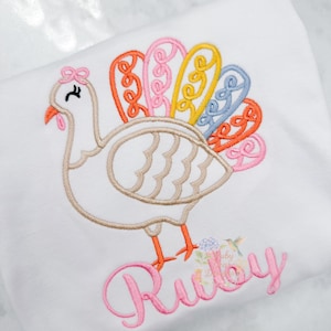 Puede incluir: Tela blanca con un diseño de pavo bordado. El pavo tiene un cuerpo beige, patas y pico naranjas, y una cola colorida con detalles en naranja, rosa, amarillo y azul. El nombre "Ruby" está bordado en rosa.