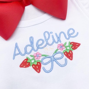 Op de afbeelding: Een witte romper met de naam "Adeline" geborduurd in blauwe cursieve letters. De naam wordt omringd door een blauwe strik en een krans van rode aardbeien en roze bloemen.