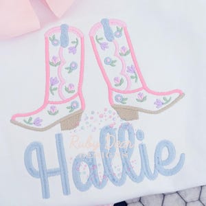 Op de afbeelding: Een wit geborduurd shirt met een paar roze en blauwe cowboylaarzen en de naam "Hallie" in blauwe cursieve letters.