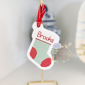 May include: A Christmas stocking ornament with the name "Brooks" embroidered in red. The stocking is white with a green body and red heel and toe. A red ribbon is attached for hanging.