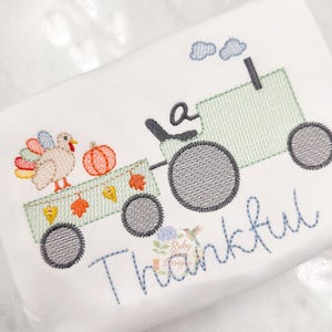 May include: Embroidered design on white fabric showing a green tractor pulling a wagon with a turkey and pumpkins. The word "Thankful" is stitched in blue script. The design includes autumn leaves and a hummingbird.