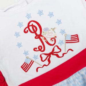 May include: A white bodice features a red embroidered script letter P, surrounded by a circle of light blue stars. Two small American flags on gold poles flank the letter, with a red ribbon bow below. A red waistband sits above a light blue gingham skirt.