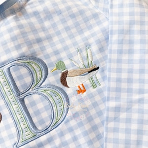 May include: A light blue gingham long-sleeve shirt with a green embroidered letter "B" and a duck with reeds embroidered on the front.