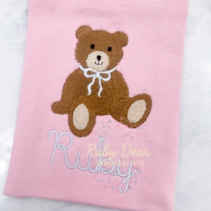 May include: A pink baby onesie with a brown embroidered teddy bear wearing a white bow. The onesie has the name "Ruby" embroidered in white cursive lettering.
