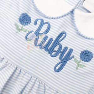 Puede incluir: Vestido a rayas azules y blancas con el nombre "Ruby" bordado en azul con escritura cursiva. El vestido presenta bordados florales azules y un cuello festoneado blanco.
