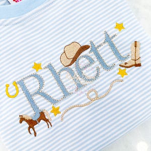 Könnte beinhalten: Ein blau-weiß gestreiftes Shirt mit dem Namen "Rhett" in blauer Stickerei, sowie einem Cowboyhut, Stiefel, Hufeisen und Lasso in Braun und Gelb.