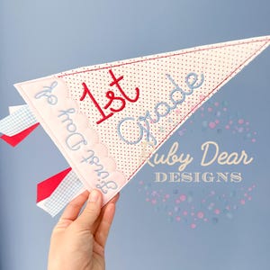 Può includere: Un gagliardetto in tessuto con le parole "1st Grade" e "First Day of" ricamate in filo rosso e blu. Il gagliardetto ha un motivo a pois ed è tenuto da una mano su uno sfondo azzurro. Il gagliardetto ha anche nastri rossi e blu.