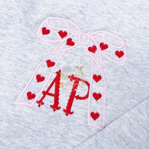 May include: A light gray fabric with a pink bow and red heart accents. The bow design frames the red embroidered letters "AP". The design also includes the text "Baby Dear Designs".