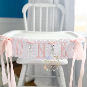 Puede incluir: Una trona blanca con una pancarta que dice "ONE" en letras rosas. La pancarta tiene un borde festoneado y cintas rosas. El fondo incluye una pared azul y una cortina blanca.