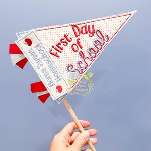 May include: A fabric pennant with the words "First Day of School" embroidered in red and blue. The pennant has a white background with red polka dots and is decorated with red ribbons and the words "Home Sweet Homeschool".