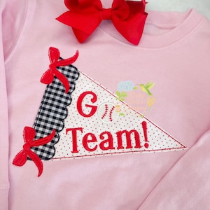 May include: A light pink sweatshirt with a red bow and a pennant design. The pennant features the words "G Team!" with a baseball graphic. The pennant has a black and white checkered pattern and red polka dots.